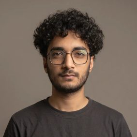 Muntaser Muttaqi, wearing glasses and a dark shirt, against a plain grey backdrop